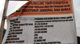 Papan informasi proyek rehabilitasi jalan dan pembangunan Jembatan Gantung Way Besai, Way Kanan, yang dikerjakan oleh Satker Pelaksanaan Jalan Nasional Wilayah II Provinsi Lampung. Proyek ini menjadi sorotan warga terkait dugaan penggunaan material batu bulat berukuran kecil pada pekerjaan pemasangan bronjong di lokasi. Foto: Tikampost.id/Marto